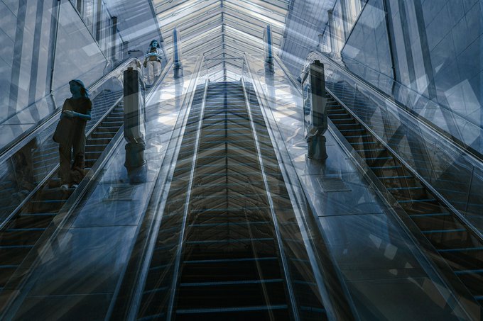 A symmetrical, mirrored image of an upward-moving escalator in a modern glass and steel building, with a person in dark clothing standing on the left side.