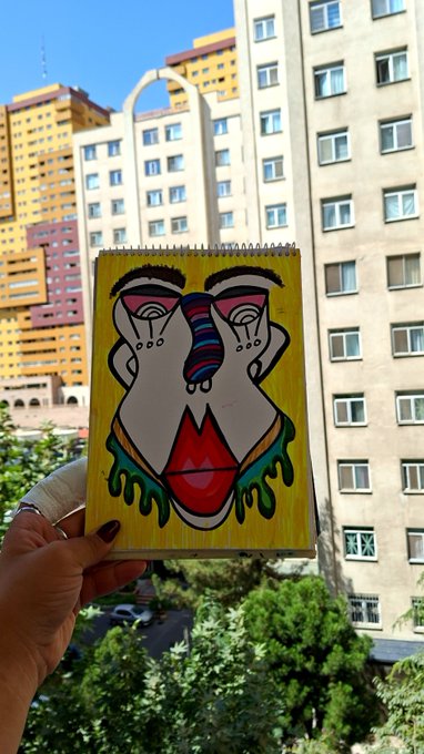 A hand holds up a notebook featuring a colorful, abstract painting of a face with dripping green accents and a red tongue, set against a backdrop of tall apartment buildings. The bold artwork pairs with the post's "50 % #SayeNFT" caption, likely promoting a half-off NFT drop inspired by the vibrant, street-art style illustration.