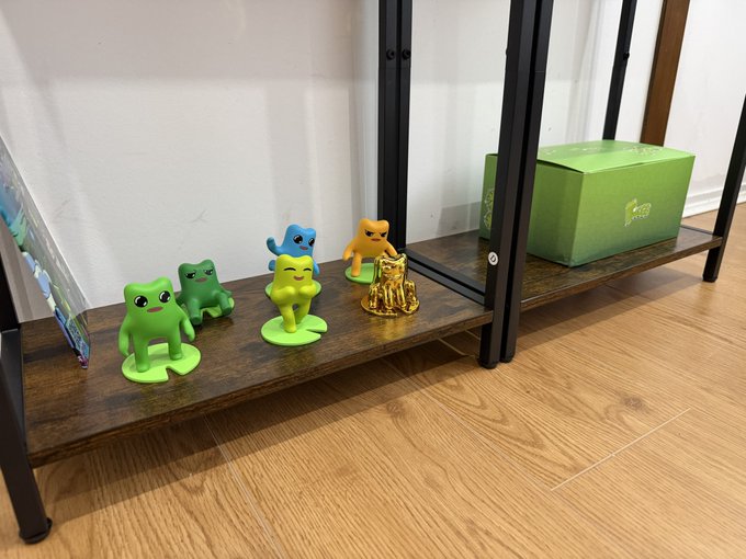 A collection of small frog figurines in green, yellow, orange, and blue colors sits on the bottom shelf of a wooden bookshelf, accompanied by a small golden figurine and a green box.