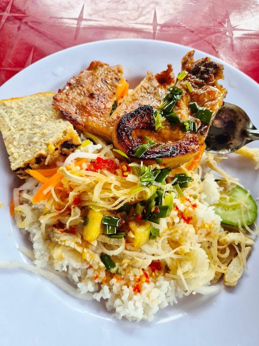 A white plate holds a Vietnamese-style dish featuring grilled fish over rice with shredded vegetables, herbs, chili, and a spoon resting on the side.