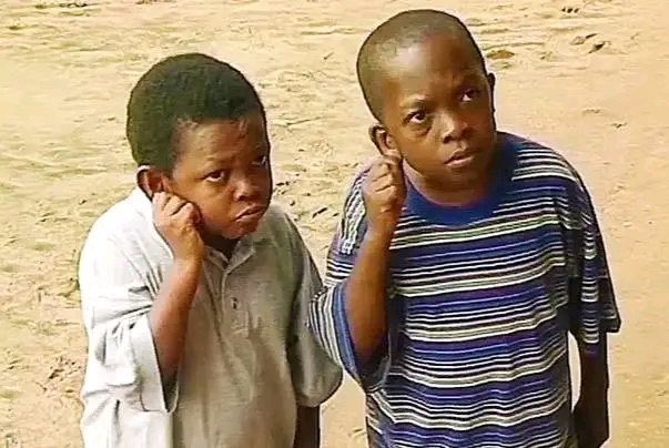 Two young boys stand on a dirt ground with puzzled and contemplative expressions, one resting his hand on his cheek while the other scratches his head.
