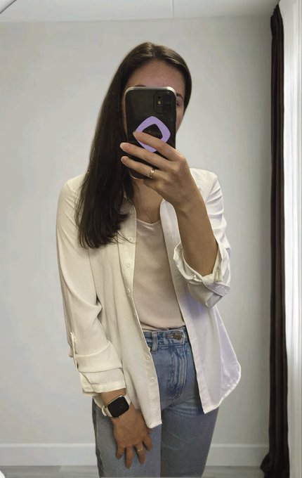 A mirror selfie of a woman with long dark hair taking a photo with her smartphone, wearing a white button-up shirt over a beige top and blue jeans, with an Apple Watch on her wrist.