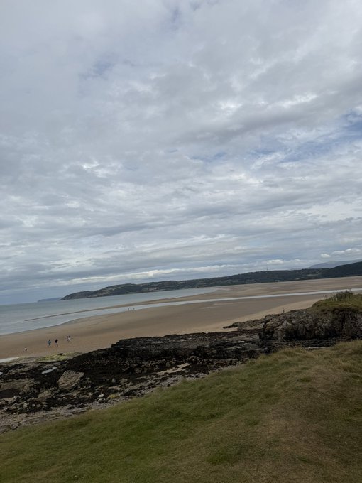 A wide sandy beach extends along a coastline under an overcast sky, with rocky outcroppings and a grassy dune in the foreground and a few people walking in the distance.