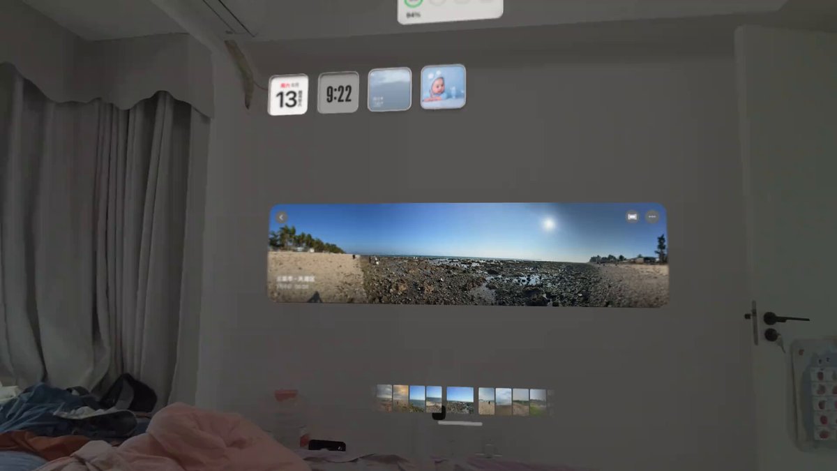 The image shows a bedroom with a large flat screen TV mounted on the wall, displaying an image of a beach scene. The room also features a bed, a window, and a door. There are two clocks visible in the room - one near the top left corner and another closer to the center right side. A remote control can be seen resting on the bed. Additionally, there is a bottle placed next to the TV screen.