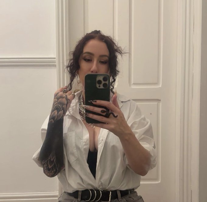 A mirror selfie of a woman with curly dark hair, a visible arm tattoo, dark nail polish, and multiple rings, wearing an unbuttoned white shirt over a black top.