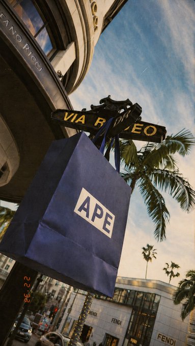 A blue "APE" shopping bag hangs from a "VIA RODEO" street sign, with FENDI store signage visible in the background of the upscale Beverly Hills shopping district, capturing the playful "Where is your bag?" reference to the $APE crypto.