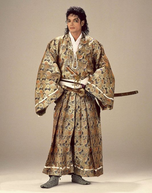 Michael Jackson poses in an ornate, gold and floral patterned traditional Japanese kimono with two swords tucked into his obi sash.