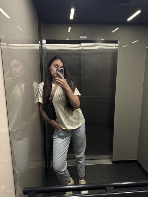 The image features a woman taking a selfie in an elevator, with her reflection visible in the mirror. She is wearing a white shirt and blue jeans, standing next to a black railing. The background of the photo shows a gray wall, which contrasts with the woman's outfit. The caption "@ethconf" suggests that this might be a social media post or a message related to an event happening at the location where she took the selfie.