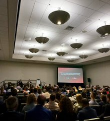 A crowded lecture hall with distinctive pendant lighting and a projection screen at the front displaying a presentation.