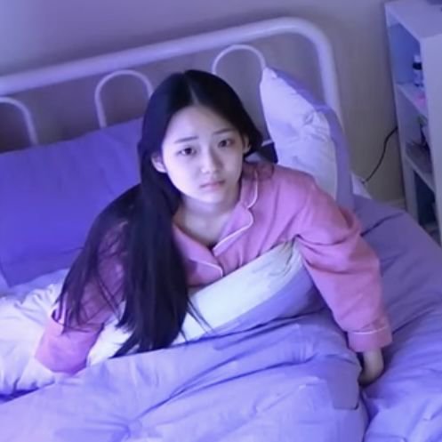 A young woman with long dark hair, wearing pink and purple pajamas, sits up in bed looking startled in a softly lit purple room.