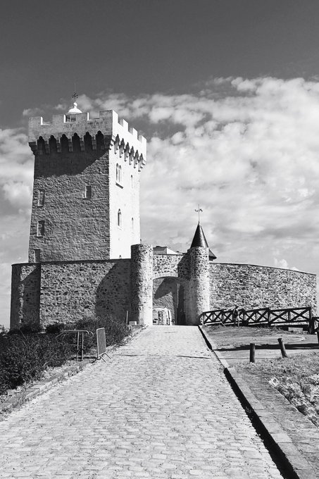 A black and white photograph features a medieval stone castle tower with battlements, accessed via a cobblestone path leading through a fortified gate.