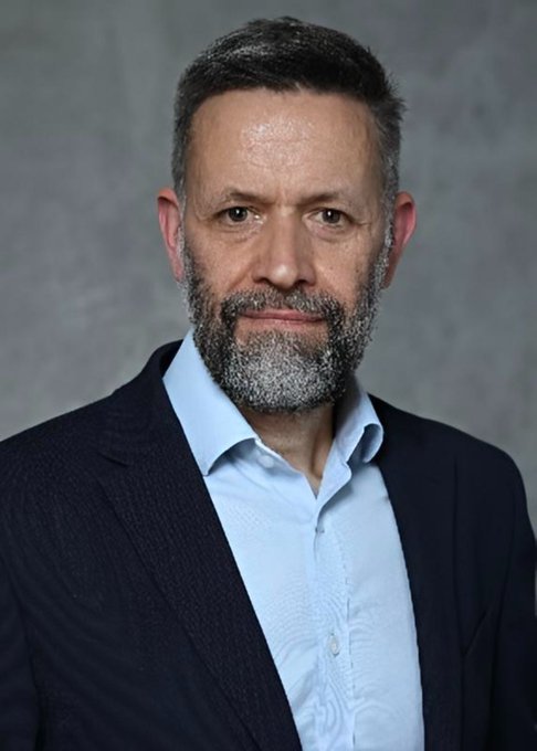 Professional headshot portrait of a middle-aged man with short dark hair and a graying beard, wearing a dark navy blazer over a light blue dress shirt against a gray background.
