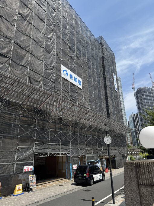 A building under construction covered in black netting and scaffolding in an urban Japanese setting, with a clock and taxi on the street.