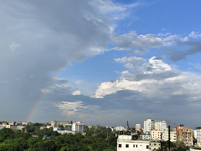 A scenic view of a cityscape under a partly cloudy sky, with a faint rainbow arcing down toward buildings and trees on the horizon.