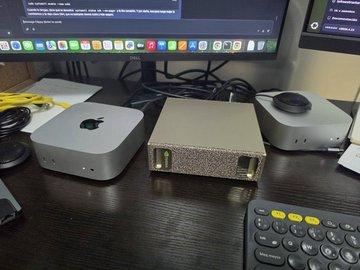 Desk setup featuring a Mac Mini, a Mac Studio, and an external device with gold corner accents in front of dual monitors, with a Stream Deck visible in the foreground.