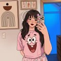 A person with long dark hair takes a mirror selfie wearing a pink shirt with a graphic design, with a blue accent wall and shelves visible in the background.