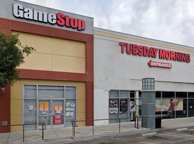 The image shows a GameStop store with its doors closed, indicating that it's not open for business at the moment. The store has a red and white sign above the entrance, which reads "GME 10Q extended deadline Tuesday 16 June after hours. Tuesday after-hours MOASS @TheRoaringKitty Psssss Psssssss".
