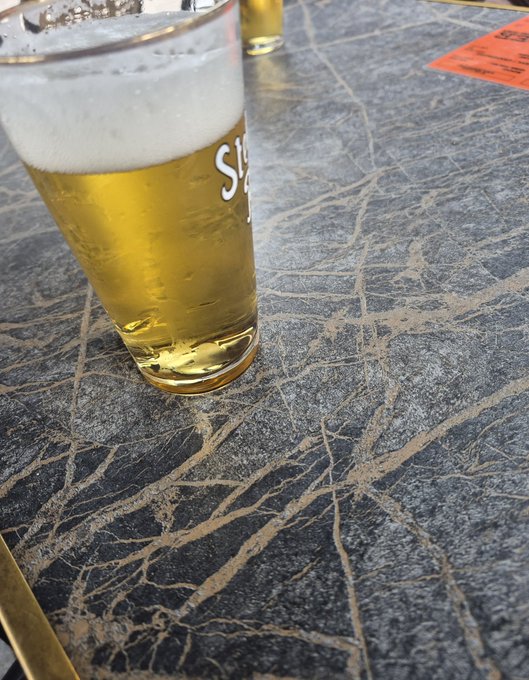 A pint glass of beer with white foam sitting on a dark marble-patterned table, with partial text 'S!' visible on the glass.