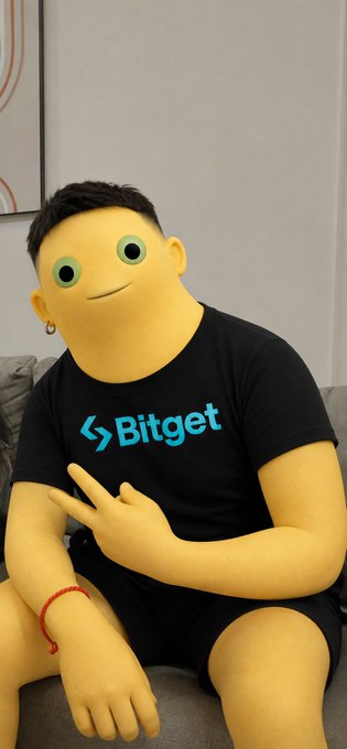 A person on a couch wears a black Bitget-branded t-shirt, but their face and hands have been digitally replaced with a cartoonish yellow emoji-like character with large green eyes.