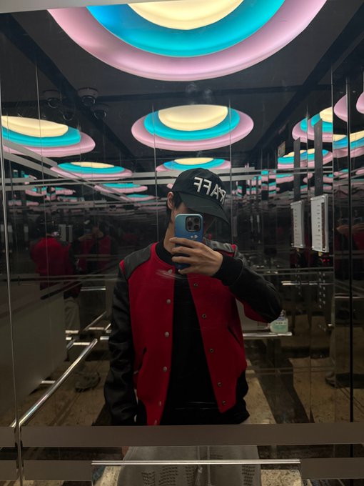 A person wearing a red and black varsity jacket and a black cap with "7L4S" text takes a mirror selfie with a blue phone in a room featuring colorful circular ceiling lights. The image aligns with the post's date (August 11, 2026) and references to LEOTAEYEONG, with "ALD1" likely relating to his album *All-Day, All Night*.