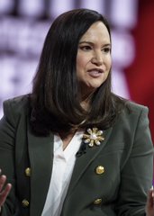 A woman with dark shoulder-length hair wearing a dark blazer with gold buttons and a brooch, speaking at what appears to be a public event with a red, white, and blue backdrop.