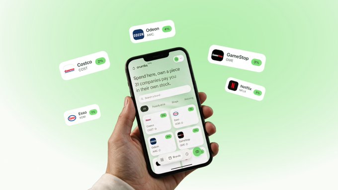 A hand holds a smartphone displaying the Crumbs app interface, which features the text "Spend here, own a piece" alongside a list of 31 partner companies with "+3%" tags, including Costco (COST), Esso (XOM), Odeon (AMC), GameStop (GME), and Netflix (NFLX). Floating cards around the phone show the same brand logos with their stock tickers, visually reinforcing the message that everyday spending can now earn onchain rewards through the platform.