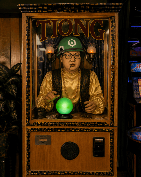 A man wearing a green cap and glasses smokes a cigar while using his finger over a glowing green crystal ball inside a fortune-telling booth labeled 'TIONG'.