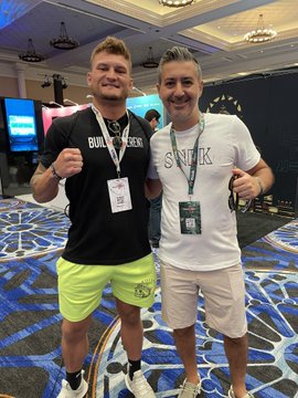 Two men pose together at what appears to be an esports event; one wears a black "BUILD A QUEEN" t-shirt with bright yellow shorts, while the other wears a white "SWIK" t-shirt with light shorts, both wearing event lanyards.