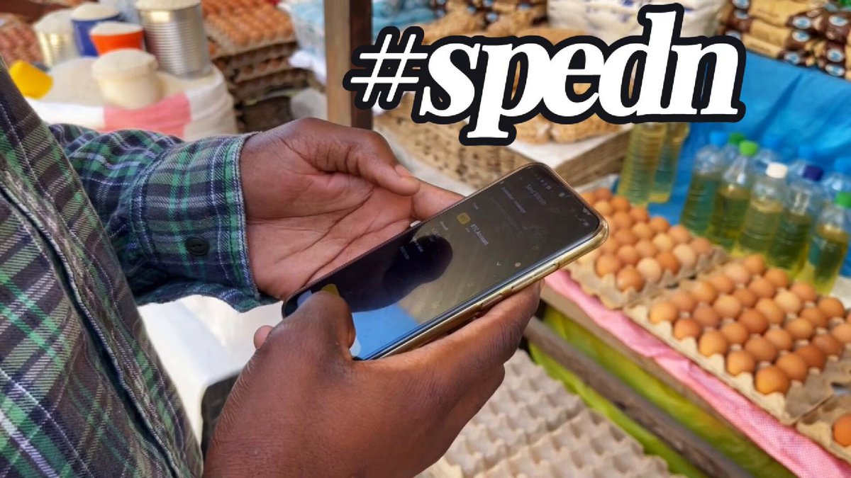 The image shows a person holding a smartphone displaying the word "SPEDN" written on it. They are standing near a table filled with eggs, suggesting they might be shopping for groceries or possibly discussing cryptocurrency. The text "Buying groceries with Bitcoin in Livingstone, Zambia" is also present in the image, indicating that this could be an advertisement or promotional material related to Bitcoin and its use as a means of payment in certain countries.