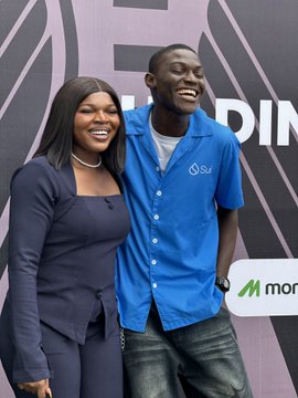 Two people smiling together, a woman in a dark blue outfit and a man wearing a blue button-down shirt with "SL2" on it