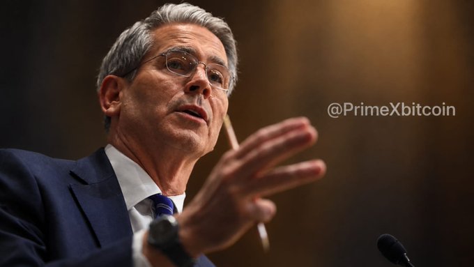 A man with gray hair and glasses wearing a navy blue suit speaks with his hand raised in a gesturing motion, with the text "@PrimeXbitcoin" visible on the right side.