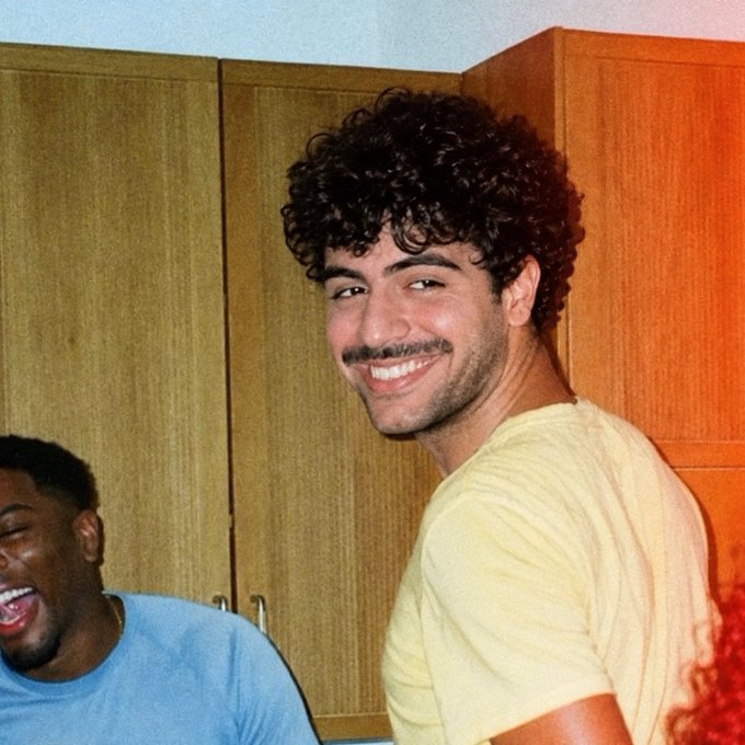 A young man with curly hair and a mustache smiles at the camera while wearing a yellow shirt, with another person laughing in the background.