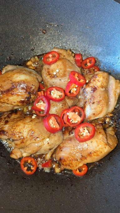 A skillet of cooked chicken thighs in sauce topped with sliced red chili peppers.