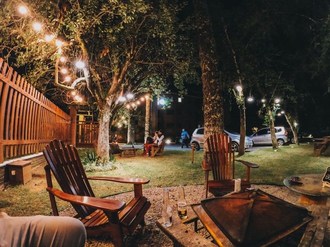 A cozy outdoor evening scene with Adirondack chairs around a fire pit, string lights in the trees, and a bottle of beer on a table.
