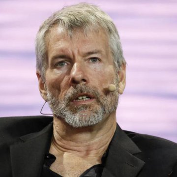 The image features a man wearing a black suit jacket with a microphone attached to his ear. He has grey hair and a beard, giving him an older appearance. The background of the image is purple, which contrasts with the man's dark attire. The caption below the photo reads "THE BIGGEST BITCOIN TREASURY COMPANY ON EARTH MAY BE ENTERING ITS MOST DANGEROUS PHASE YET UNDERSTANDING THE RISKS OF THE BIGGEST BITCOIN TRADING COMPANY". This suggests that the man is discussing a potential threat to Bitcoin trading companies, possibly due to market conditions or other factors.