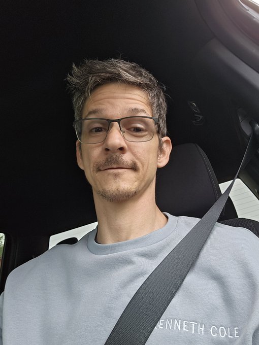 A selfie of a man wearing glasses and a light blue Kenneth Cole t-shirt, seated in a car with his seatbelt fastened.