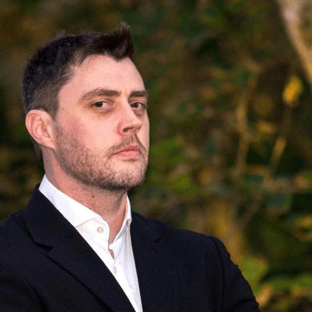 Portrait of Andre Cronje, founder of Flying Tulip, shown in a dark suit against a blurred outdoor background. The image accompanies his view that DeFi has largely evolved into on-chain finance because many current protocols lack true decentralization.