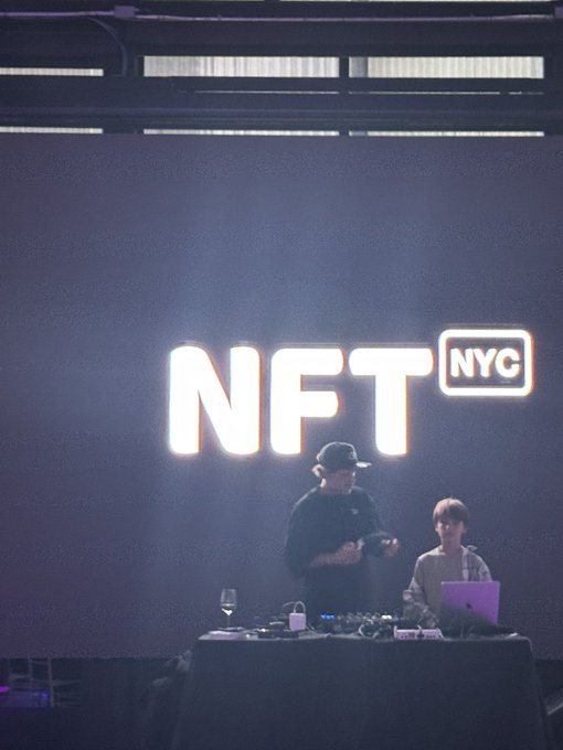 Two people stand behind a DJ setup on stage with a large illuminated 'NFT NYC' sign displayed in the background.