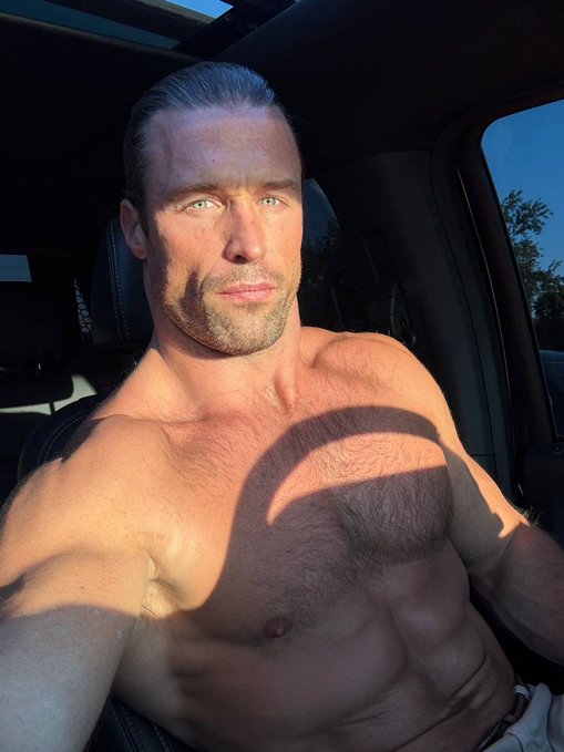 A shirtless, muscular man takes a selfie in the driver's seat of a car, with sunlight casting a shadow across his chest.