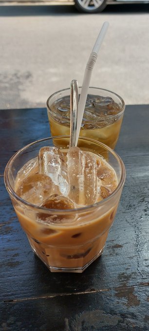 Two glasses of iced coffee with milk and straws sit on a dark wooden table, with the blurred street and a car wheel visible in the background.