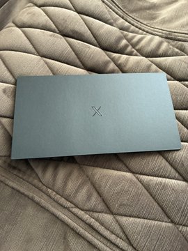 A sleek dark gray envelope or card sleeve featuring a minimalist "X" logo rests on a tan quilted fabric surface, likely containing the new X Money card referenced in the post about its cash back and APY benefits.