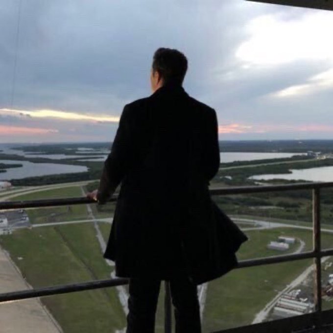 A man in a black coat stands on a high balcony looking out over a sprawling landscape of lakes and fields at sunset.