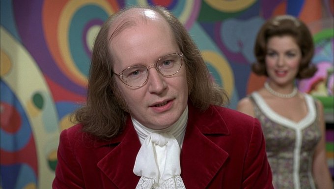 The image features a man with long hair wearing glasses, a red coat, and a white tie standing next to another woman who has short brown hair and is also dressed up for an event. The background of the image shows a colorful mural or wall art behind them. The caption "Ironwood... Ms Ironwood $ZEC" suggests that this scene might be from a movie or TV show where the main characters are named Ironwood and Ms Ironwood, possibly indicating their roles in the story.