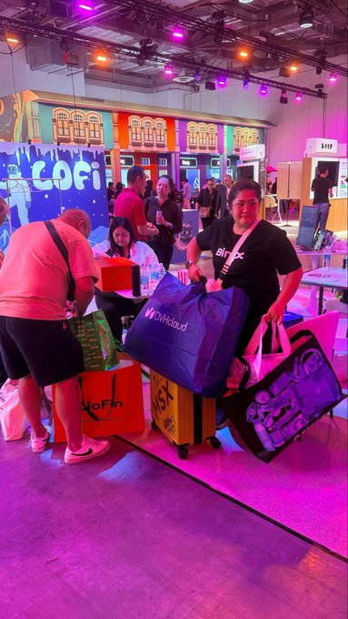 Several women smile in the foreground at a vibrant event, posing with bags including a blue 'Offcloud' duffel, a yellow 'MSX' suitcase, and a pink gaming PC case, with a colorful 'CoFi' booth behind them.