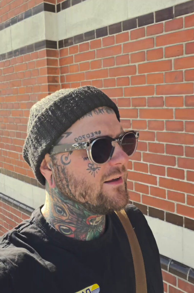 The image features a man wearing sunglasses, a black hat, and has tattoos on his neck and face. He appears to be posing for a photo or selfie with a brick wall as the backdrop. The caption of the image reads "It'S official @SolOnChain website going live tonight. ... Launchpad ... Marketplace ... NFT Raffles All in 1 stop for all your NFT'S needs." This suggests that the man is promoting an online marketplace, possibly related to NFTs or digital art, and he wants to share his experience with others by taking a photo of himself in front of the brick wall.
