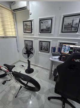 An indoor room with white walls displaying multiple framed black-and-white photos, a standing fan, an air conditioner, a desk with items, a gaming chair, and a bicycle leaning in the foreground.