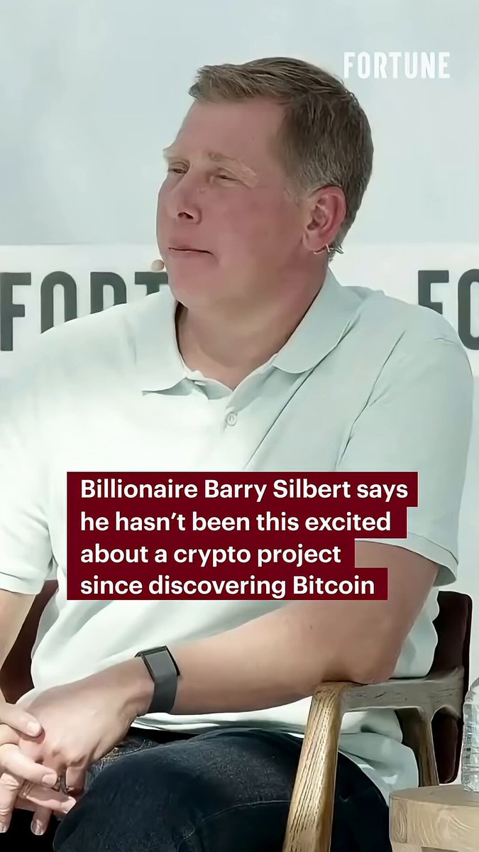 The image features a man sitting down, wearing a white shirt with his hands folded in front of him. He appears to be engaged in conversation or listening to someone speaking. The background includes a sign that reads "Fortune" and another sign that says "Bitcoin".