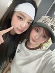 A selfie of two young women; one wears a white headband and makes a peace sign, while the other wears a cap and a gold chain, both posing close together.