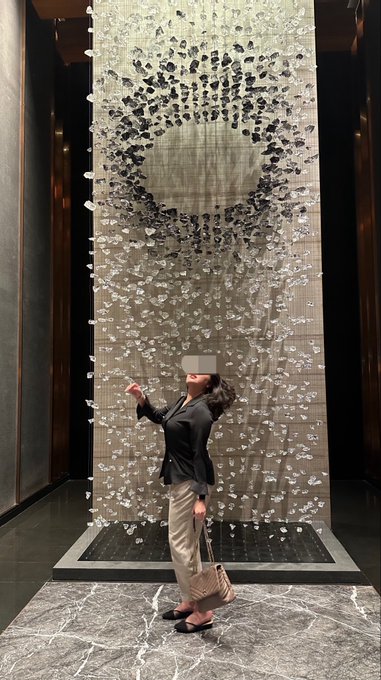 A woman stands in profile looking up at a large, illuminated art installation of a circular wire mesh sculpture with hanging crystals, located in a modern building with marble flooring.