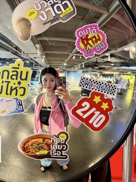 A young woman takes a mirror selfie in what appears to be an arcade or entertainment venue, wearing a pink jacket and holding a phone, with a large bowl of food in the foreground. Colorful Thai promotional stickers overlay the image, including "HYPE 21.10," "2/10," and "เพิ่ม 20," which playfully tie into her post about studying crypto tokens like $HYPE and $ONDO while celebrating her DeFi earnings with a themed meal.